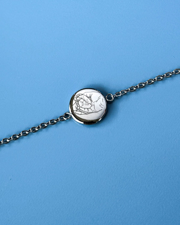 IBI PORTRAIT armbånd i sølvfarvet rustfrit stål med stregtegning af hænder graveret på rund plade, mod blå baggrund