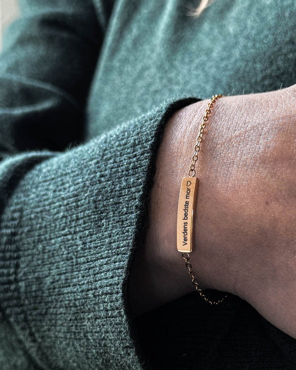 EVI armbånd i guldbelagt stål med "Verdens bedste mor" ♡ graveret på aflang plade, båret om håndleddet til grøn strik
