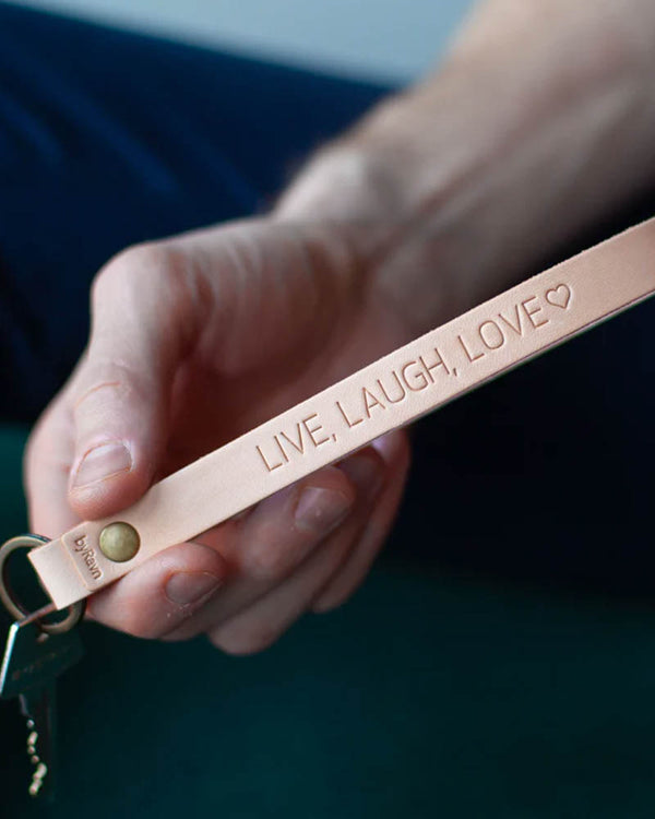 LUX nøglering i naturfarvet læder med messingnitte og personlig gravering "Live, laugh, love" ♡, holdt i hånden med nøgle