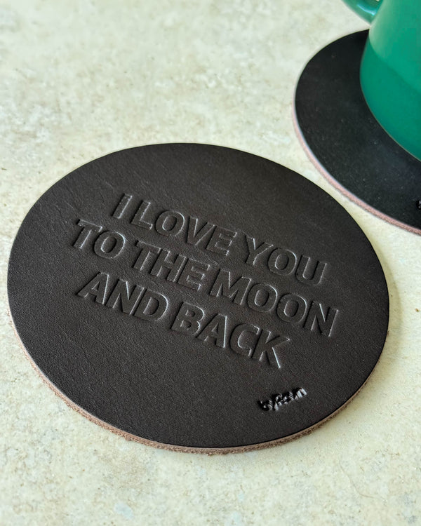 MOKKA bordskåner i mørkebrunt læder præget "I love you to the moon and back" på lyst bord ved grøn kop