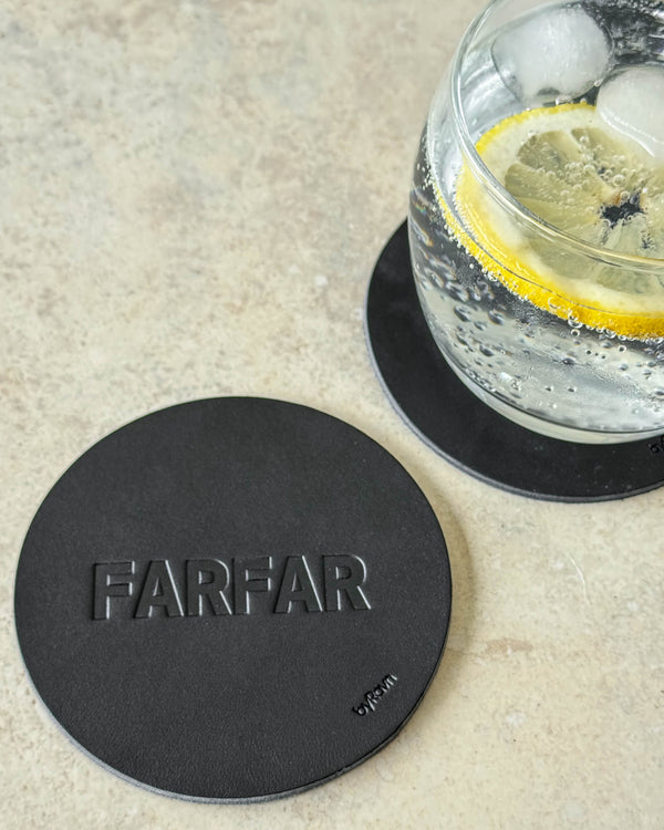 MOKKA bordskåner i sort læder præget "Farfar" på lyst bord ved glas med gin og tonic og citronskive
