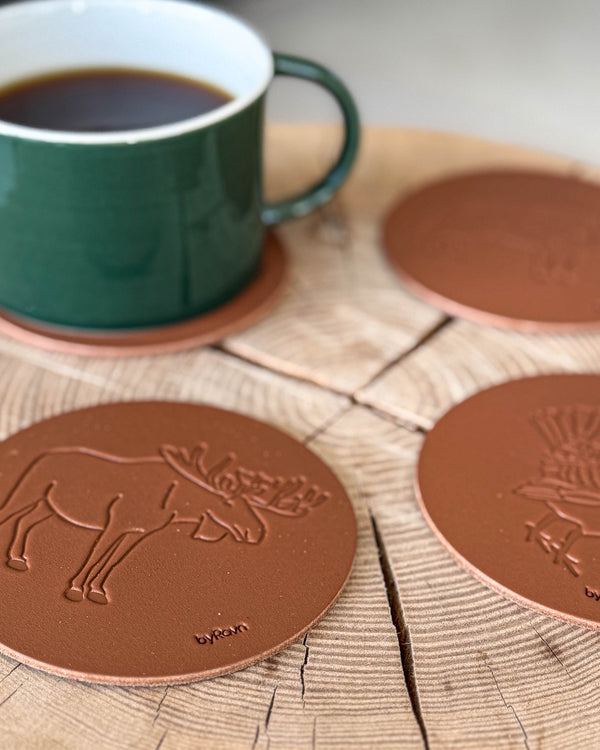 MOKKA jagt bordskånere i cognac læder præget med elg og tjur på træskive ved grøn kaffekop på coaster