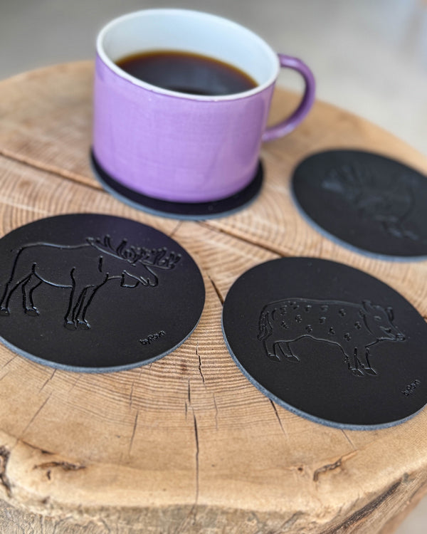 MOKKA jagt bordskånere i sort læder præget med elg, vildsvin og tjur på træskive med lilla kaffekop på coaster