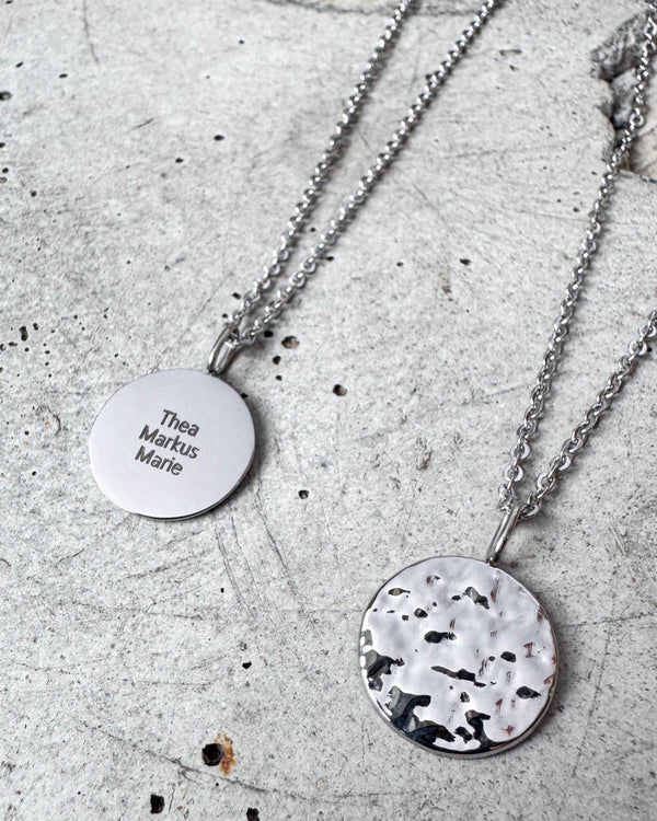 MOON QUOTE navnehalskæde i sølvfarvet rustfrit stål med navnene "Thea Markus Marie" graveret, hamret side vist ved siden af, på beton