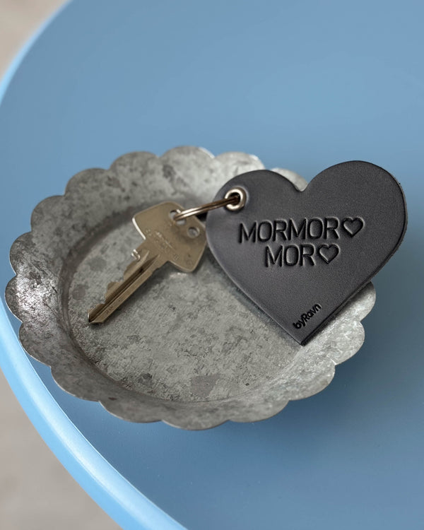 ROSE hjerte-nøglering i sort læder med graveringen "Mormor ♡ Mor ♡" og nøgle i zinkskål på blåt bord