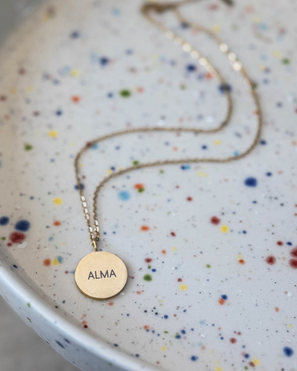 SIA NOTE navnehalskæde i guldbelagt stål med navnet "Alma" graveret på rundt vedhæng, på spættet keramikfad