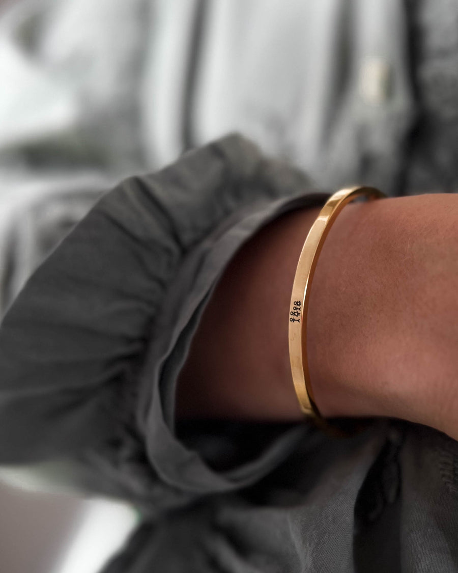 VENUS STICK armbånd i guldbelagt stål med tændstikfamilie graveret, båret om håndleddet til grå bluse