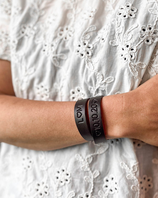 DUO læderarmbånd i mørkebrunt læder med graveringerne "L ♡ M" og "♡ 10.10.20 ♡" om håndleddet, hvid broderet bluse