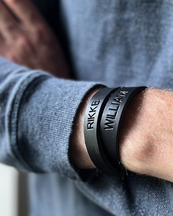 DUO læderarmbånd i sort læder med navnene "Rikke" og "William" graveret, båret om mandehåndled i grå strik