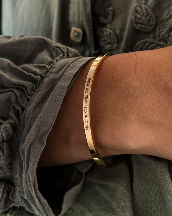 VENUS armbånd i guldbelagt stål med graveringen "Nicoline ♡ Laurits ♡ Kristian" om håndleddet, grå bluse