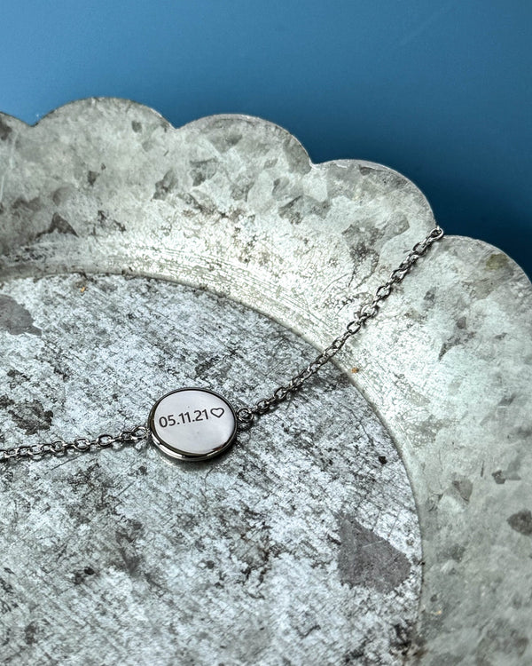 IBI NOTE armbånd i sølvfarvet rustfrit stål med "05.11.21" ♡ graveret på rund plade, på zinkfad mod blå baggrund