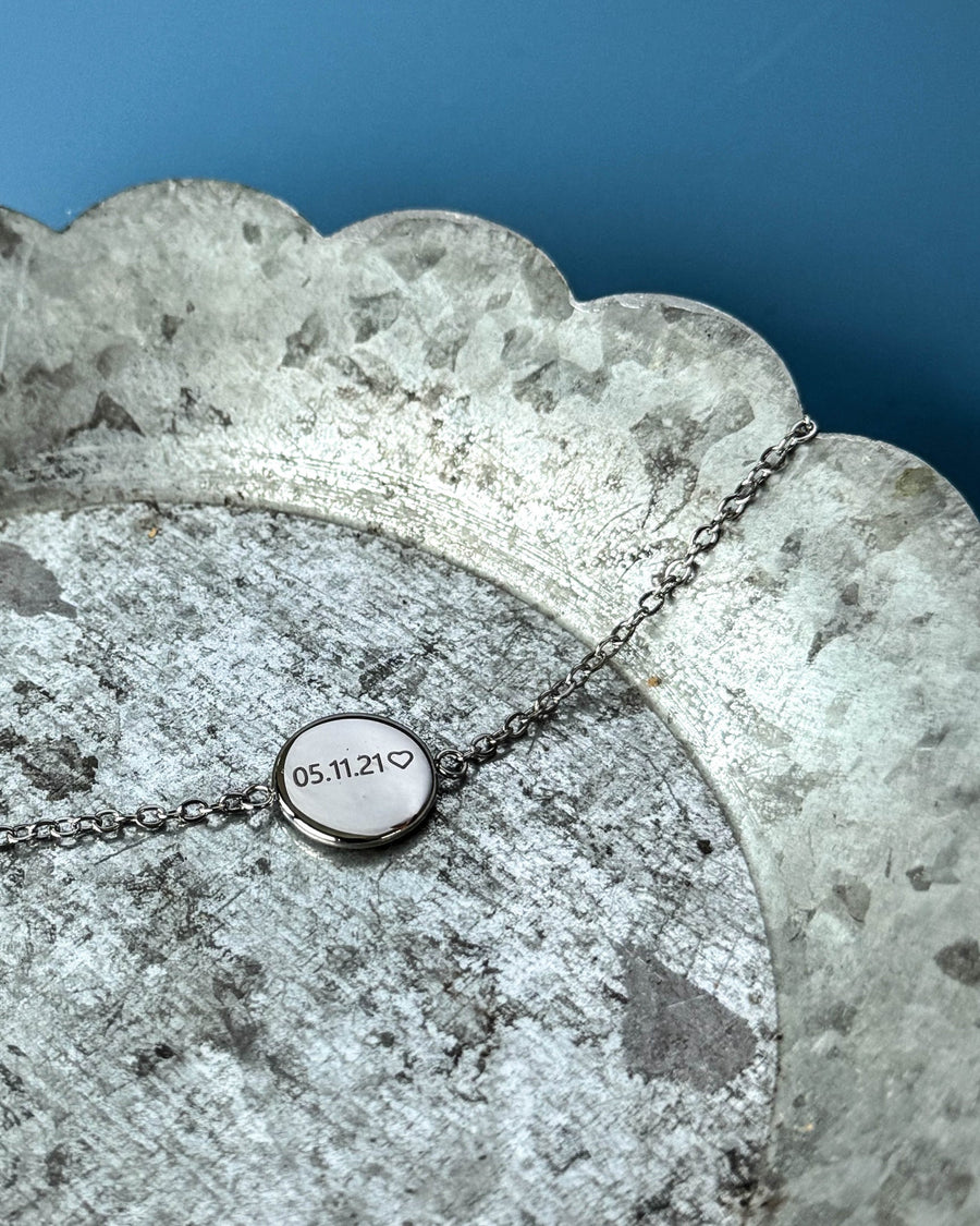 IBI NOTE armbånd i sølvfarvet rustfrit stål med "05.11.21" ♡ graveret på rund plade, på zinkfad mod blå baggrund
