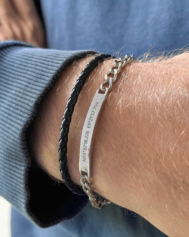 ARA armbånd i sølvfarvet rustfrit stål med koordinaterne "N55°32'18.226 E9°21'57.704" graveret, båret med sort flettet læderarmbånd