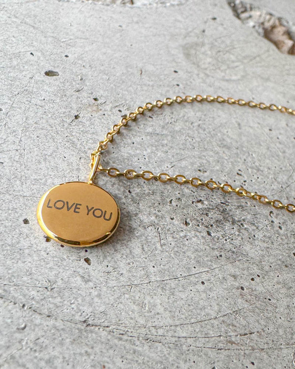 AYA NOTE halskæde i guldbelagt stål med graveringen "Love you" på lille rundt vedhæng, liggende på beton