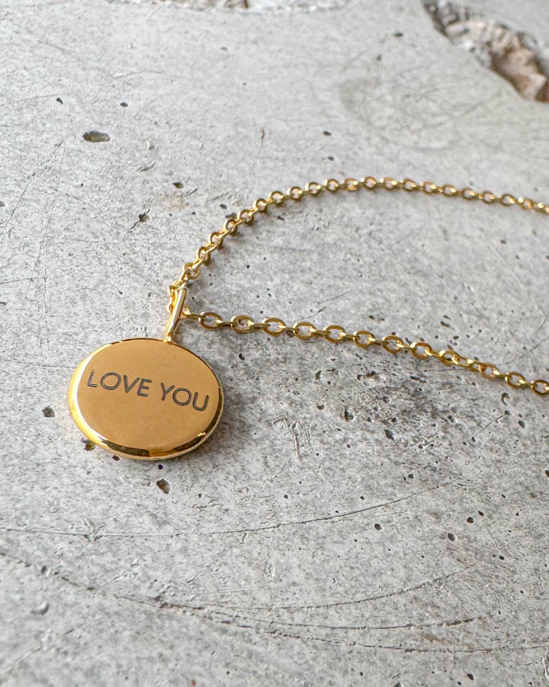 AYA NOTE halskæde i guldbelagt stål med graveringen "Love you" på lille rundt vedhæng, liggende på beton