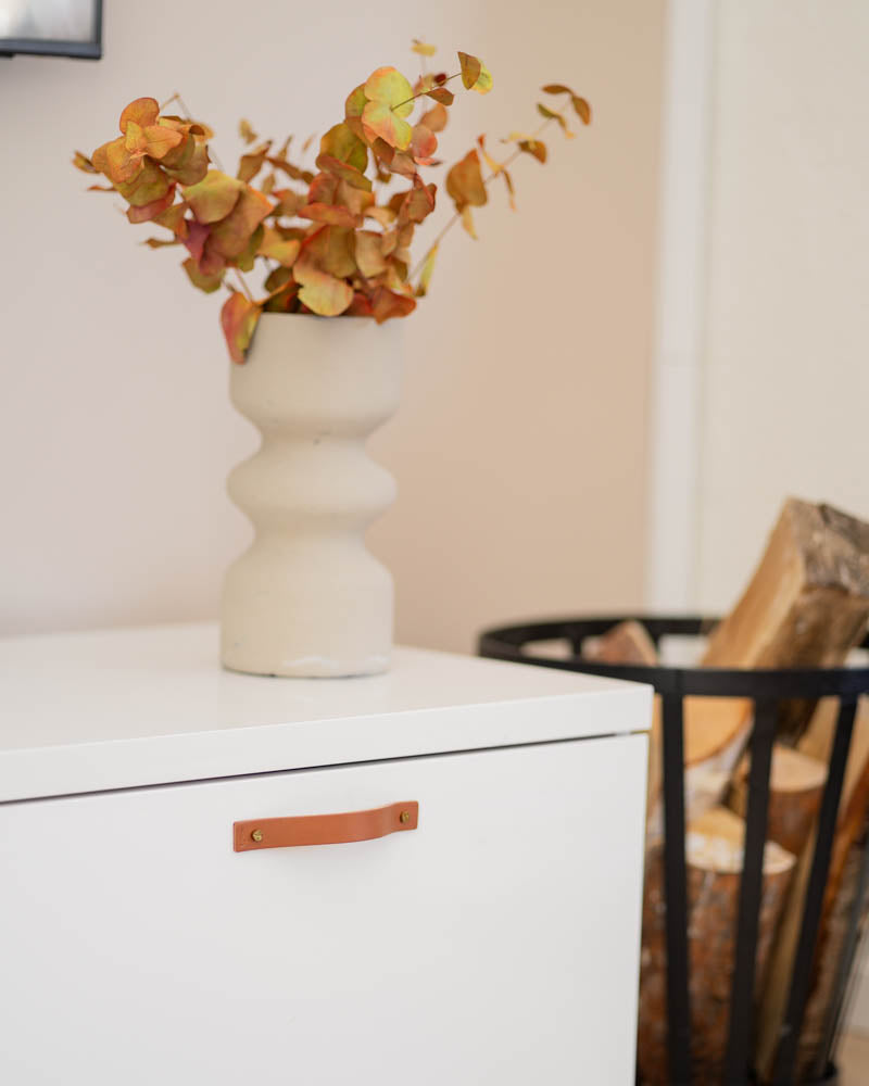 BAND greb i cognac læder på hvid kommode med beige vase med efterårsgrene og brændekurv ved siden af