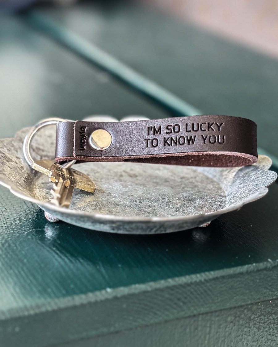 BOND nøglering i mørkebrunt læder med graveringen "I'm so lucky to know you" og nøgle i zinkfad på grønt bord