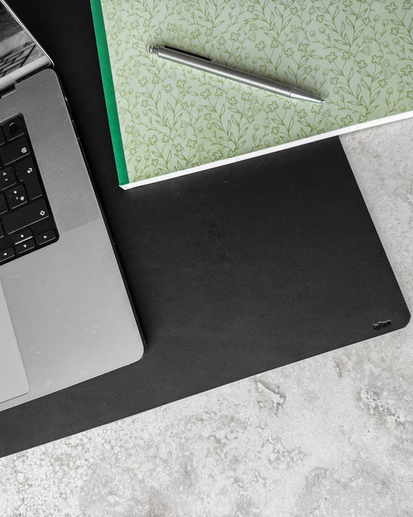 DESK skriveunderlag i sort læder set oppefra med laptop, blomstret notesbog og kuglepen på lys bordplade