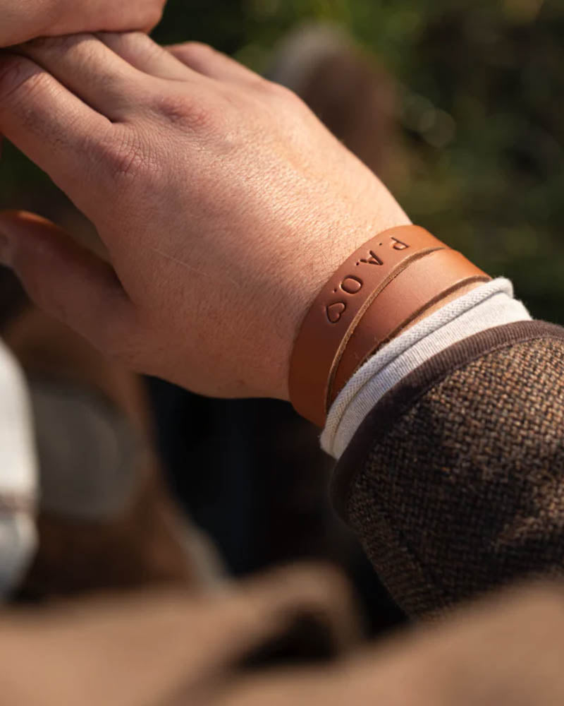 DUO læderarmbånd i cognac læder med initialerne "P.A.O." ♡ graveret, båret om håndleddet udendørs i solskin