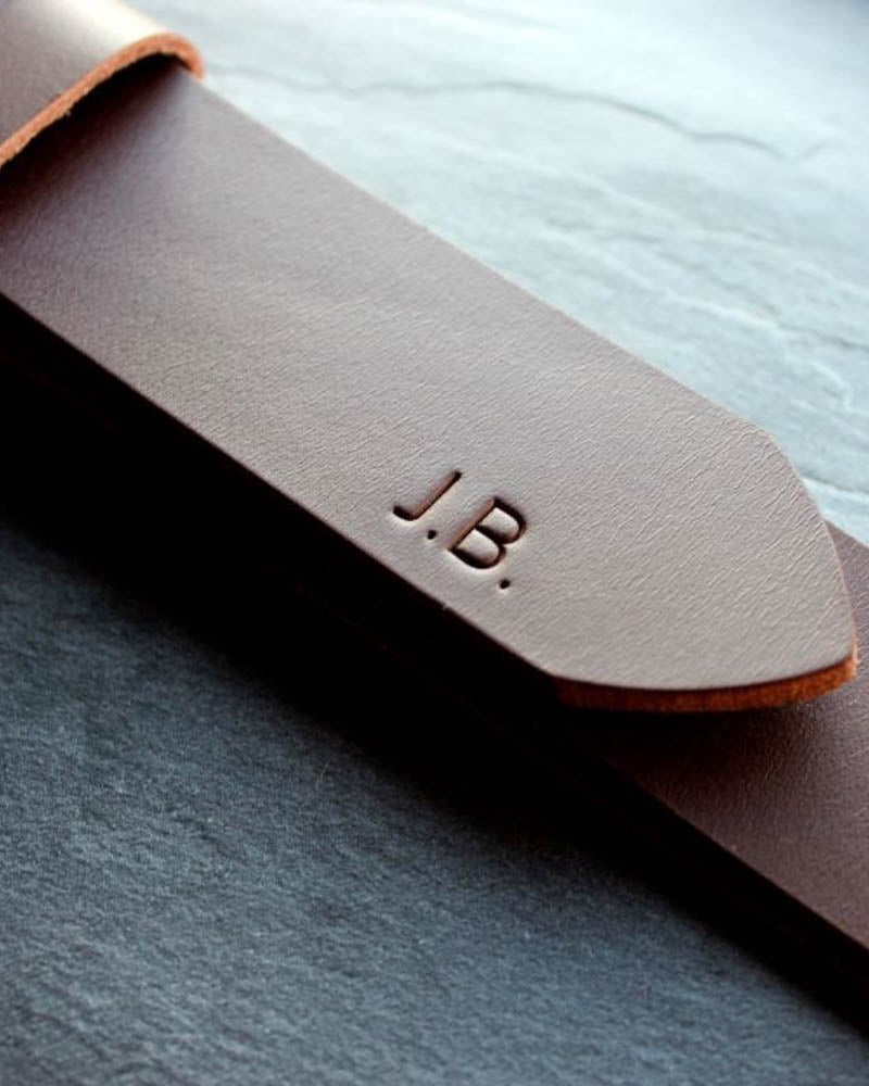 EDGE bælte i mørkebrunt læder set tæt på med initialerne "J.B." graveret på spidsen, på skiferplade