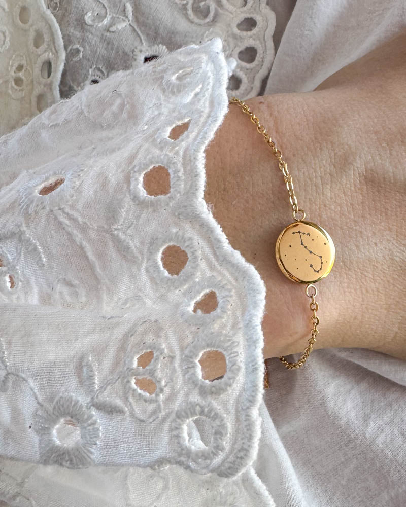 IBI STAR armbånd i guldbelagt stål med stjernebillede graveret på rund plade, båret om håndleddet til hvid blondebluse