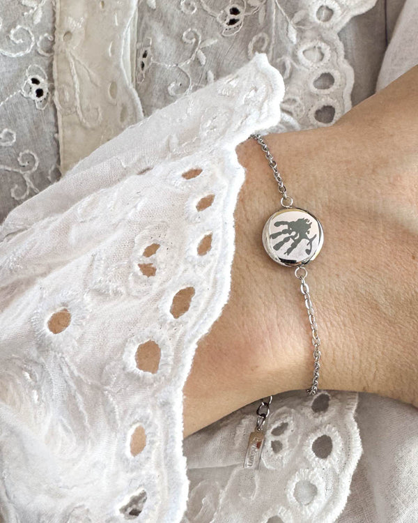 IBI MOTIF armbånd i sølvfarvet rustfrit stål med graveret håndaftryk, båret om håndleddet til hvid blondebluse