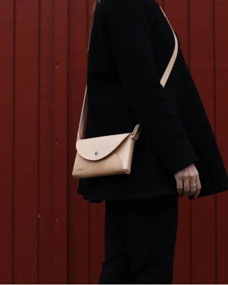 INA skuldertaske i naturfarvet læder båret crossbody af kvinde i sort tøj foran rød træbeklædning