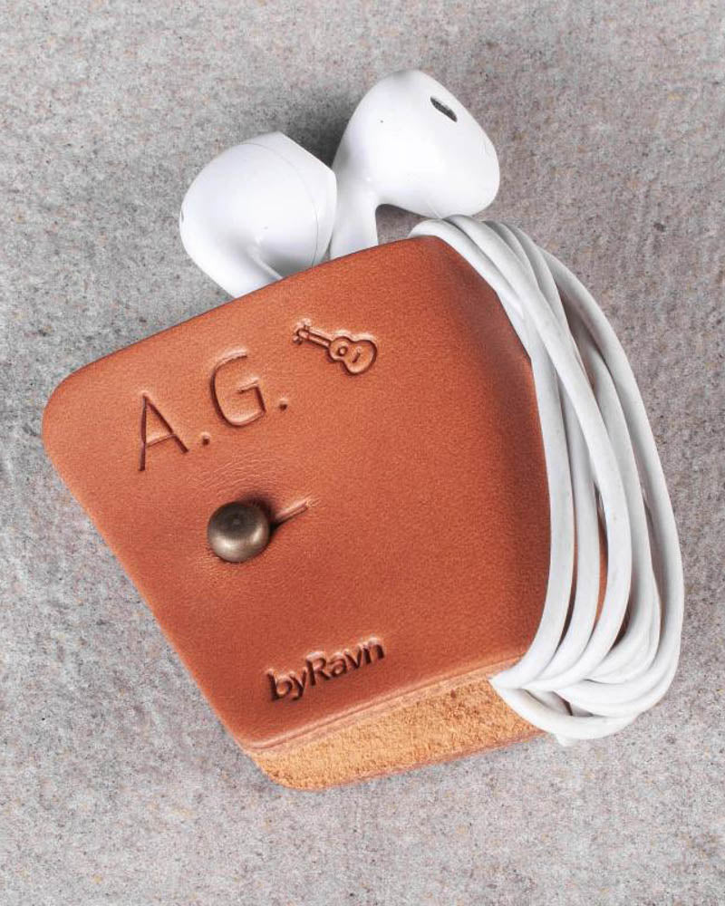 KAX høretelefonholder i cognac læder med initialerne "A.G." og guitar graveret, med earpods på grå sten