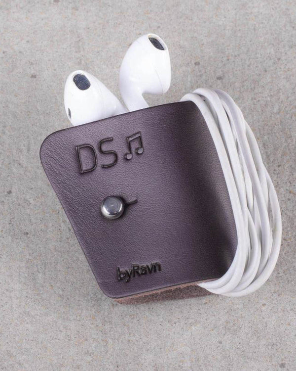 KAX høretelefon-clips i mørkebrunt læder med hvide earpods viklet om og graveringen "DS" med node, på betonbaggrund
