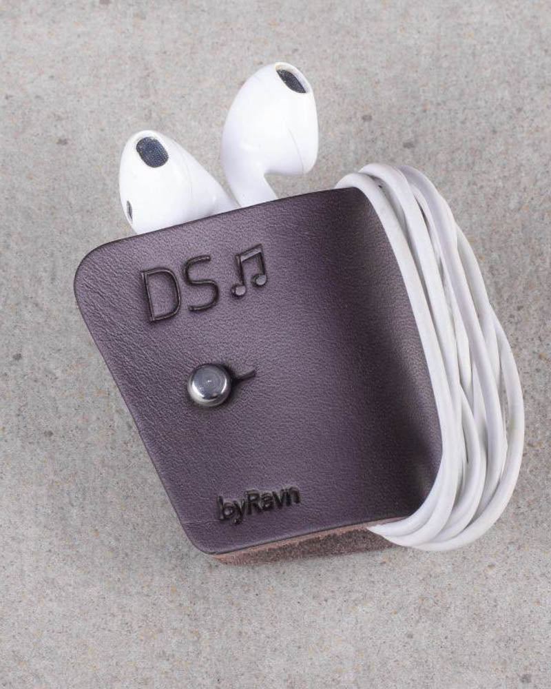 KAX høretelefon-clips i mørkebrunt læder med hvide earpods viklet om og graveringen "DS" med node, på betonbaggrund