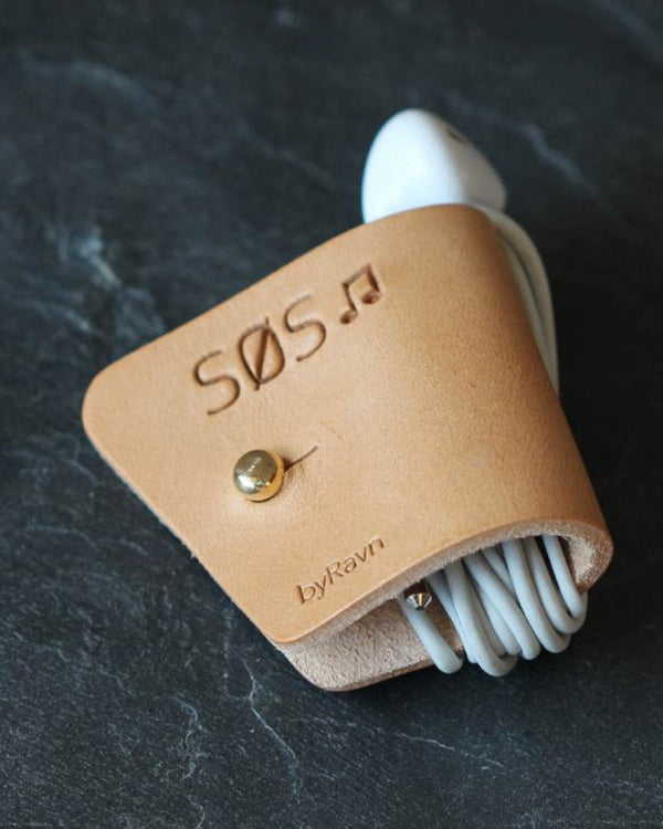 KAX høretelefonholder i naturfarvet læder med graveringen "Søs" ♪ og messingknap, med earpods på mørk skifer