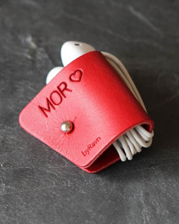 KAX høretelefon-clips i rødt læder med graveringen "Mor" ♡ og hvide høretelefoner viklet om, på mørk skifer