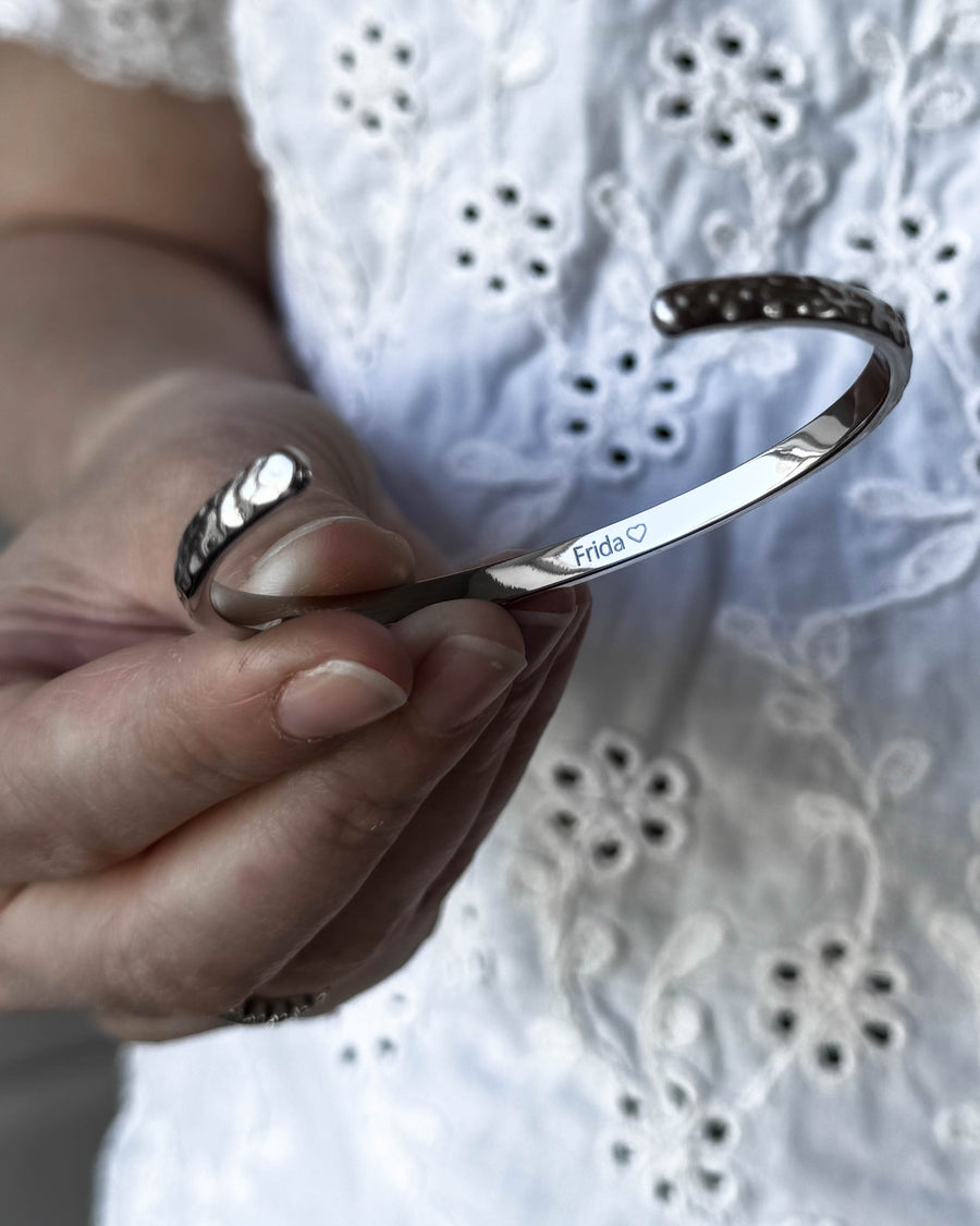 LUNA armbånd i sølvfarvet rustfrit stål holdt i hånden, med navnet "Frida" ♡ graveret på indersiden