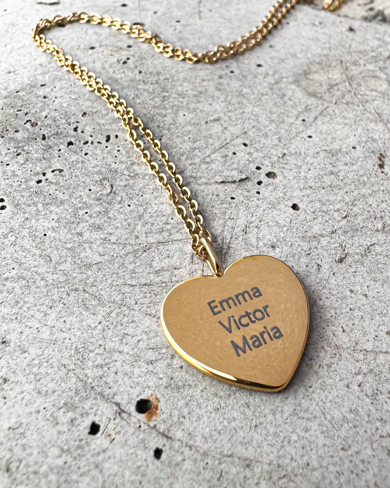 LUV QUOTE navnehalskæde i guldbelagt stål med navnene "Emma Victor Maria" graveret på hjertevedhæng, liggende på beton