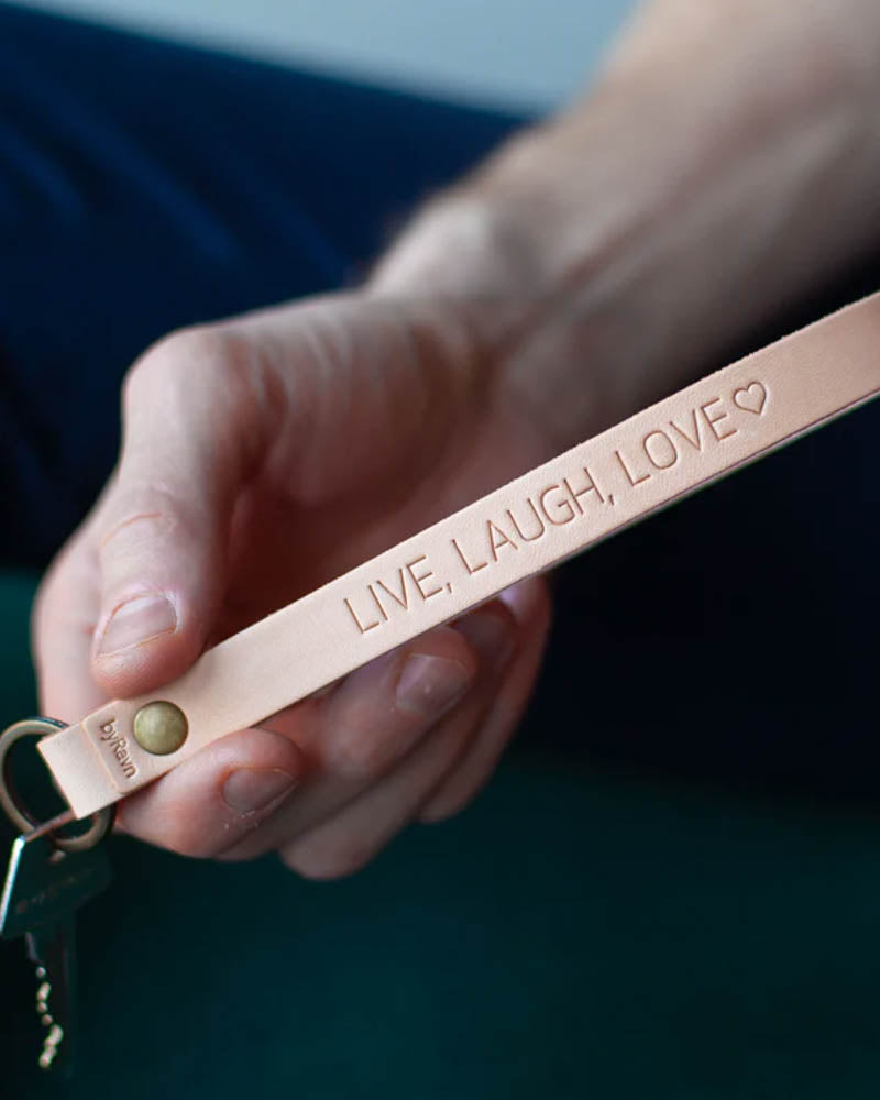 LUX nøglering i naturfarvet læder med messingnitte og personlig gravering "Live, laugh, love" ♡, holdt i hånden med nøgle