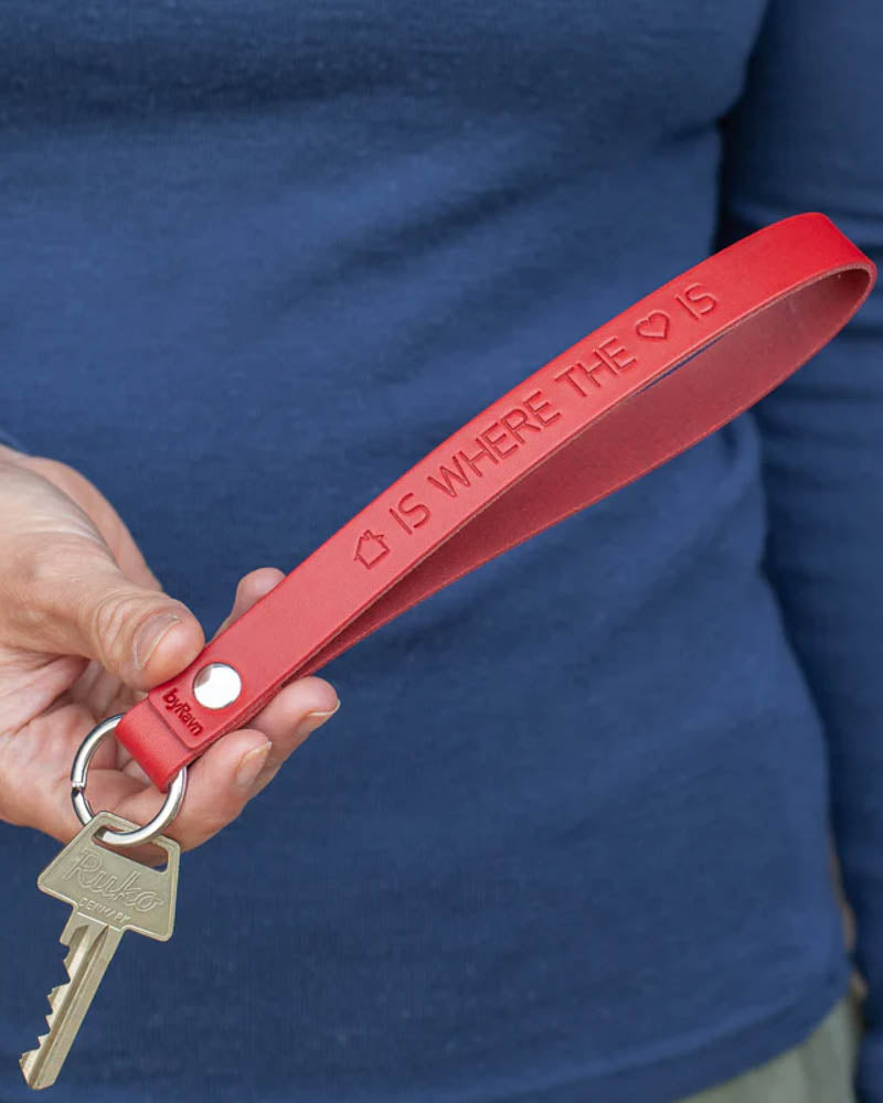 LUX nøglering i rødt læder med graveringen "Home is where the heart is" skrevet med hus- og hjertesymbol, holdt i hånden