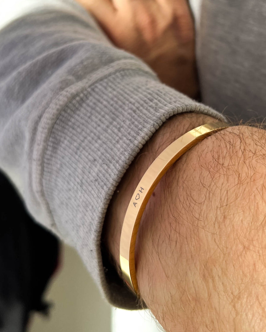 MARS armbånd i guldbelagt stål med initialerne "A ♡ H" graveret om mandehåndled, grå sweatshirt