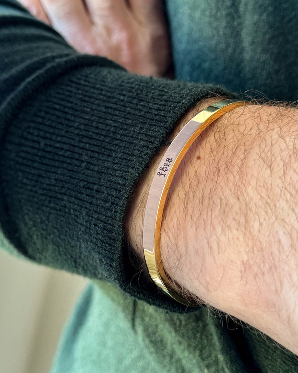 MARS STICK armbånd i guldbelagt stål med tændstikfamilie graveret, båret om håndleddet til mørkegrøn strik