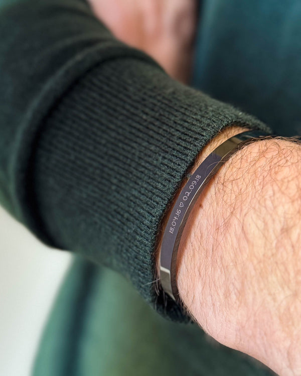 MARS SCRIPT armbånd i sort stål med datoerne "18.04.16 ♡ 02.09.18" graveret i håndskrift, båret til mørkegrøn strik