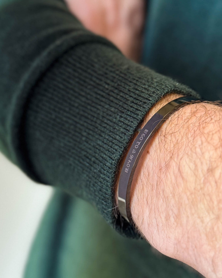 MARS SCRIPT armbånd i sort stål med datoerne "18.04.16 ♡ 02.09.18" graveret i håndskrift, båret til mørkegrøn strik