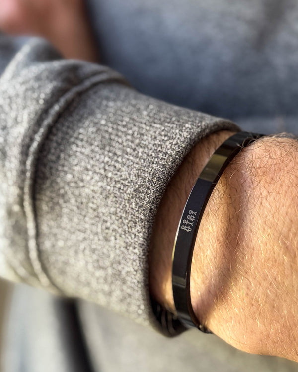 MARS STICK armbånd i sort stål med tændstikfamilie graveret, båret om håndleddet til grå strik