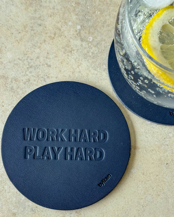 MOKKA bordskåner i mørkeblåt læder præget "Work hard play hard" på lyst bord ved glas med citronskive
