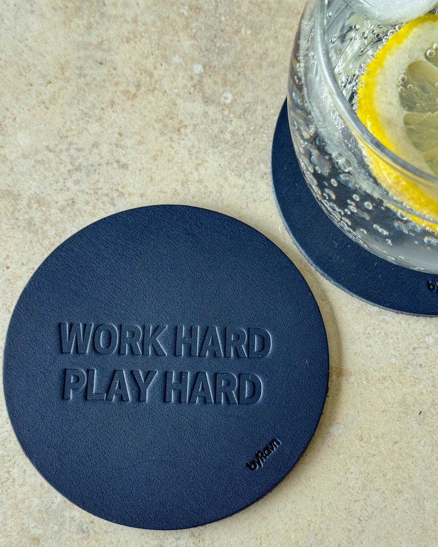 MOKKA bordskåner i mørkeblåt læder præget "Work hard play hard" på lyst bord ved glas med citronskive