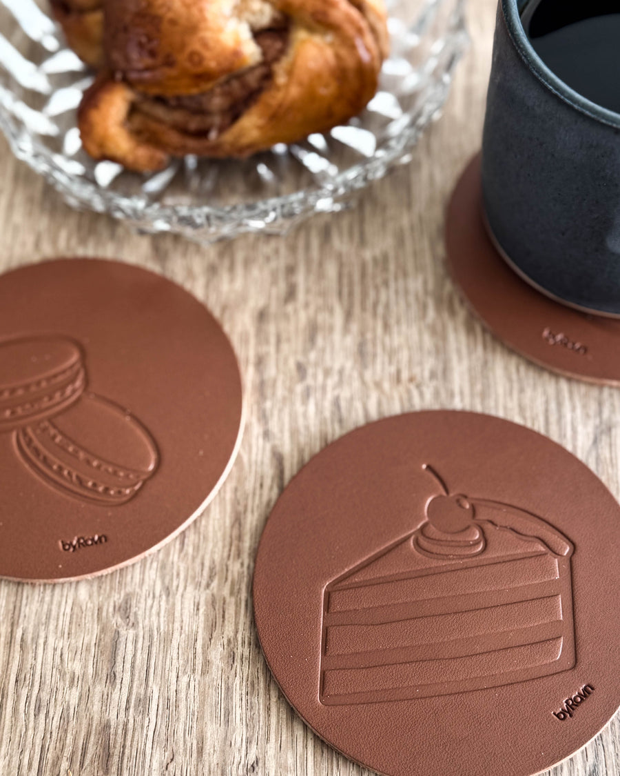 MOKKA kaffe & kage bordskånere i cognac læder præget med lagkage og macarons på træbord ved kanelsnegl og sort kop