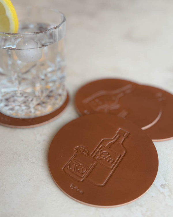 MOKKA spiritus bordskånere i cognac læder præget med ginflaske og drink, på lys stenflade ved glas med citronskive