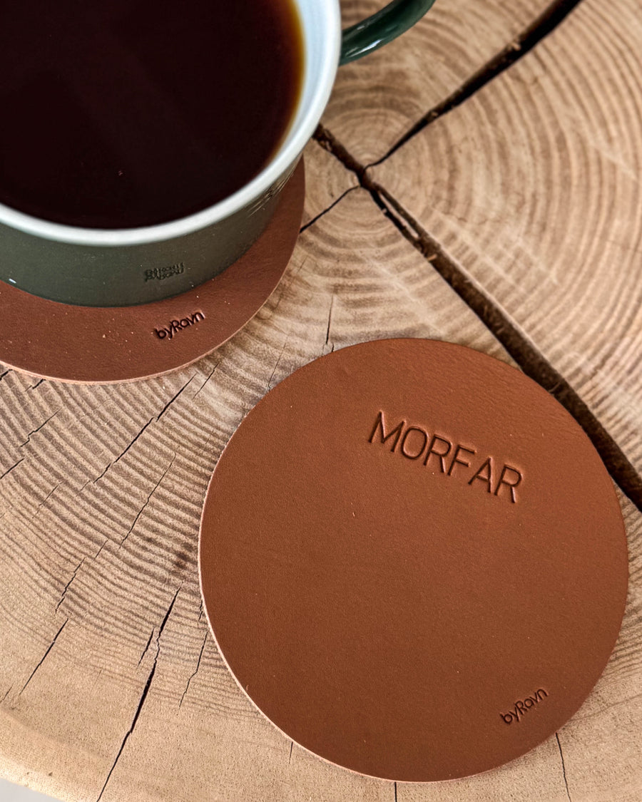 MOKKA rund bordskåner i cognac læder med graveringen "Morfar" på træskive ved siden af grøn kaffekop på coaster