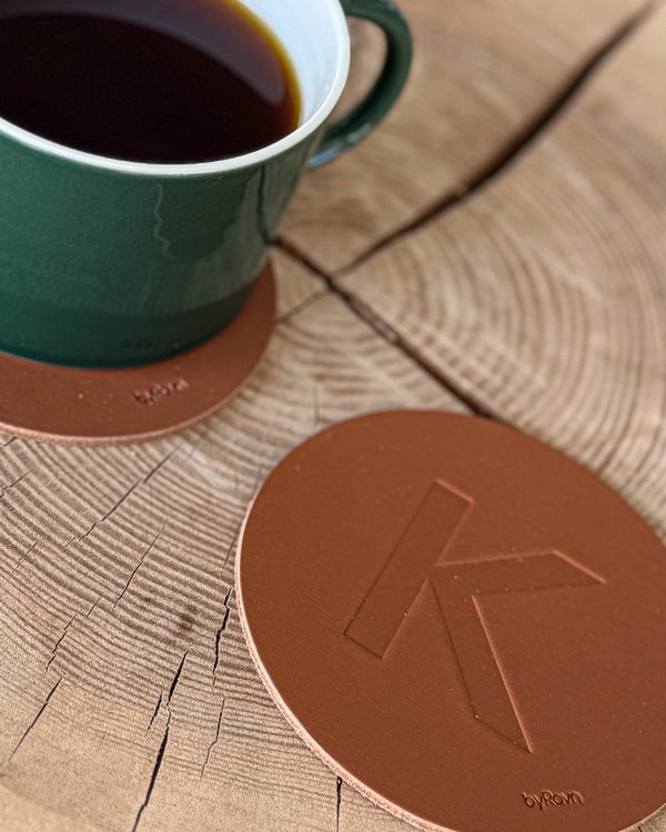 MOKKA bordskåner i cognac læder præget med bogstavet "K" på træskive ved grøn kaffekop på coaster