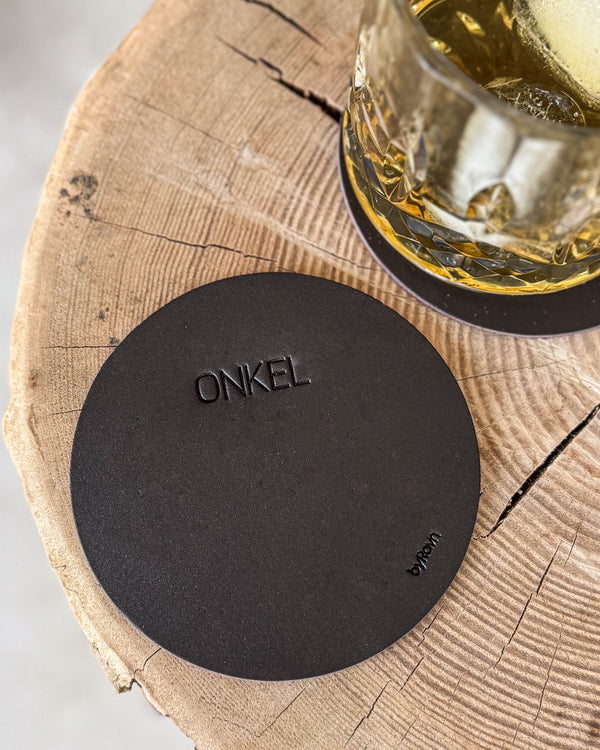 MOKKA rund bordskåner i mørkebrunt læder med graveringen "Onkel" på træskive, whiskyglas på coaster bagved