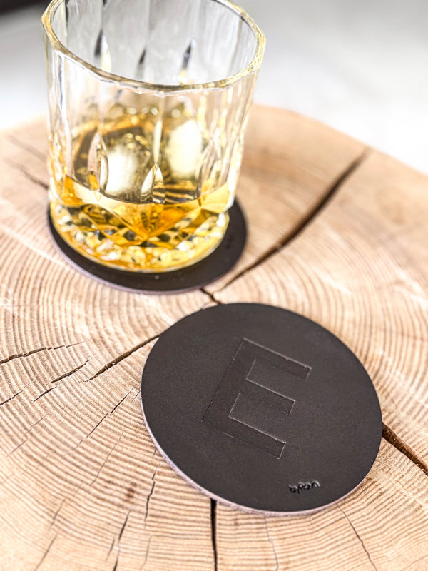 MOKKA bordskåner i mørkebrunt læder præget med bogstavet "E" på træskive ved whiskyglas på coaster