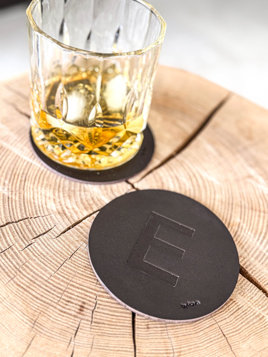MOKKA bordskåner i mørkebrunt læder præget med bogstavet "E" på træskive ved whiskyglas på coaster